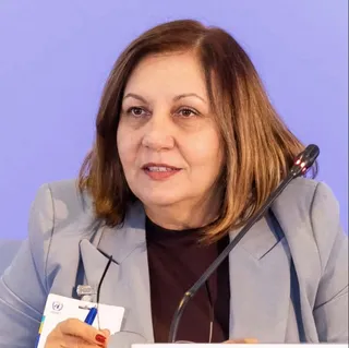 O femeie cu părul castaniu, purtând un sacou gri și un tricou închis la culoare, stă în fața unui fundal violet lângă un microfon negru.