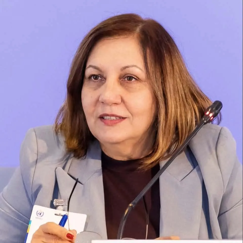 O femeie cu părul castaniu, purtând un sacou gri și un tricou închis la culoare, stă în fața unui fundal violet lângă un microfon negru.