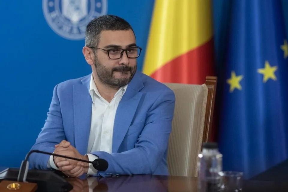 Un bărbat cu ochelari și sacou albastru stă la o masă în fața unor steaguri.