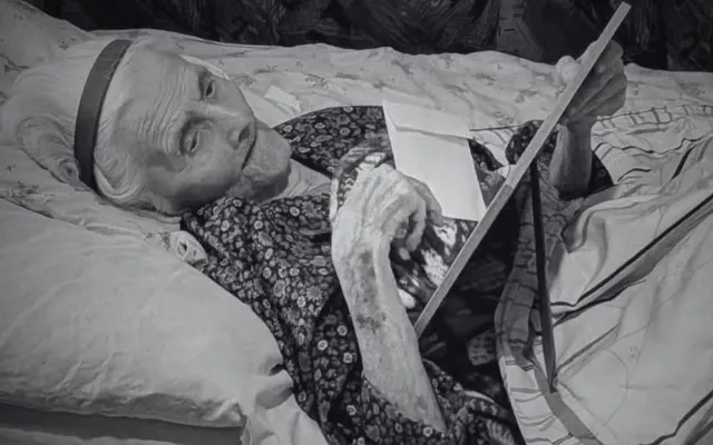 O femeie în vârstă, cu părul alb, poartă o bentiță și o rochie cu imprimeu floral, stând întinsă în pat și ținând o carte.