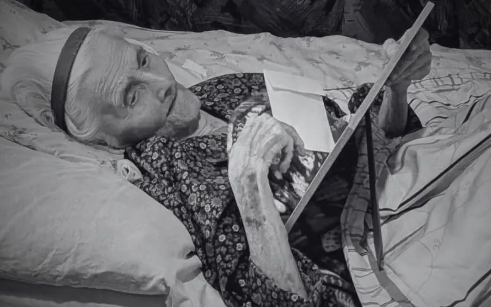 O femeie în vârstă, cu părul alb, poartă o bentiță și o rochie cu imprimeu floral, stând întinsă în pat și ținând o carte.