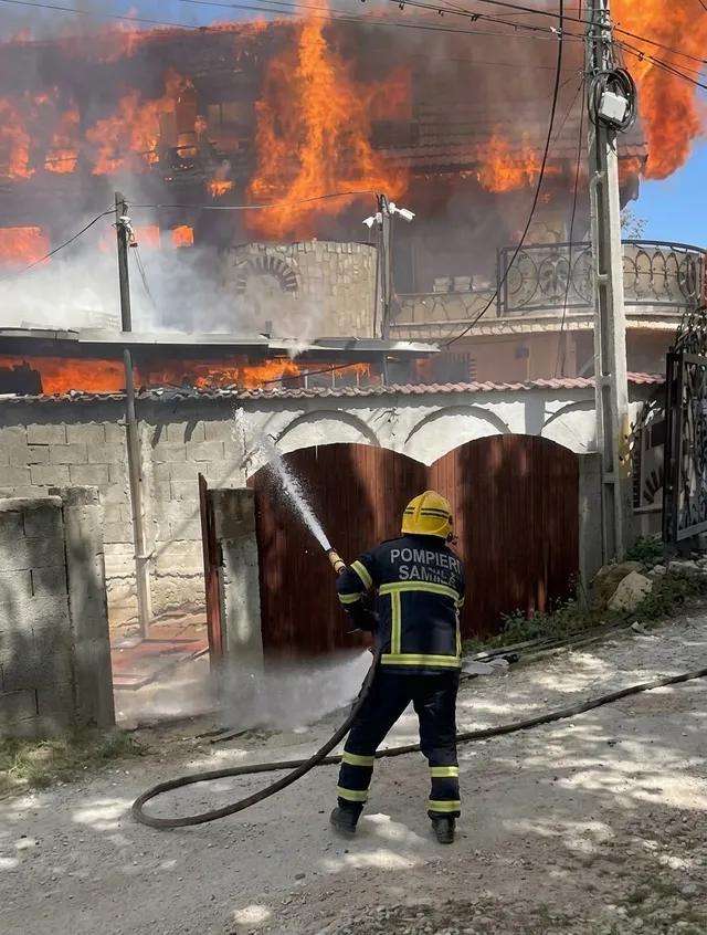 Un pompier în echipament de protecție folosește un furtun pentru a stinge flăcările care ies de peste un zid și o poartă maro.