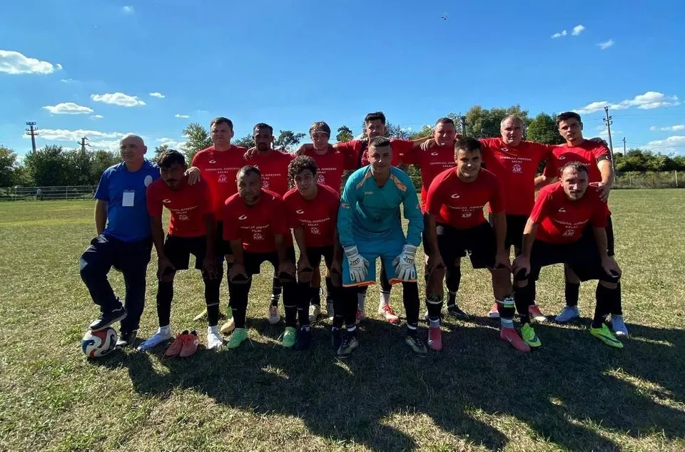Un grup de bărbați îmbrăcați în echipamente sportive roșii și unul în echipament de portar albastru pozează pe un teren de fotbal sub cerul senin.