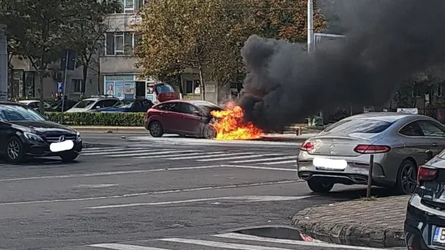 Un autoturism roșu este în flăcări pe un drum asfaltat, emanând un nor dens de fum negru în apropierea unui zebru.