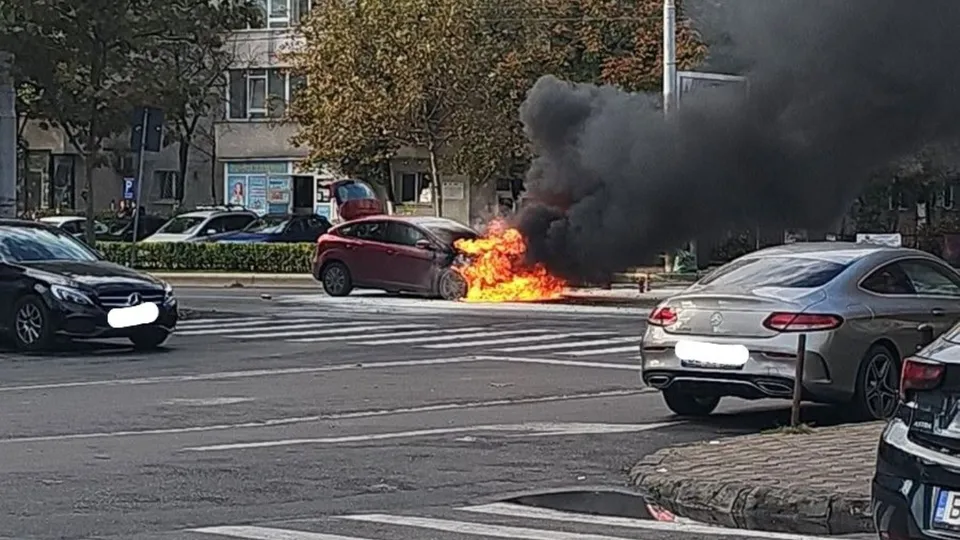 Un autoturism roșu este în flăcări pe un drum asfaltat, emanând un nor dens de fum negru în apropierea unui zebru.