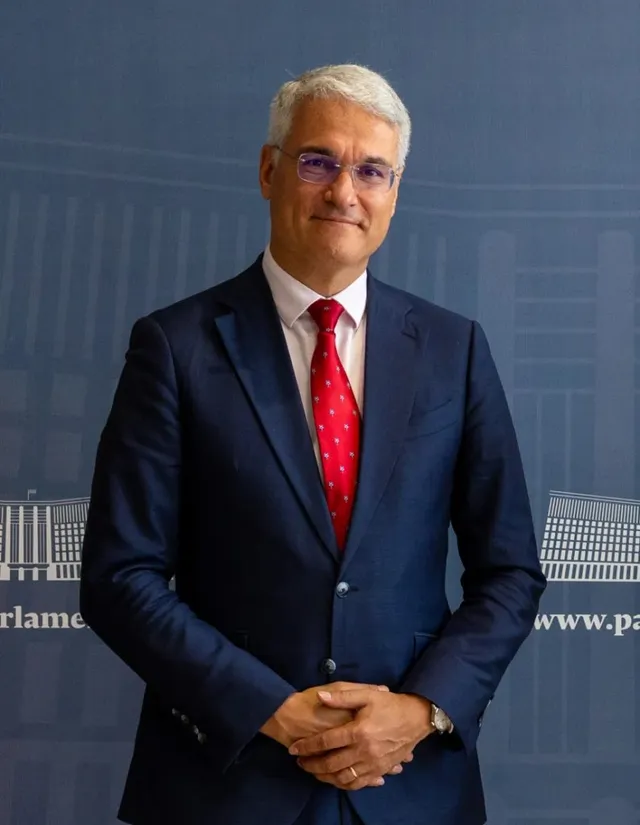 Un bărbat cu părul gri, purtând ochelari, sacou albastru, cămașă albă și cravată roșie, stă cu mâinile încrucișate în față.
