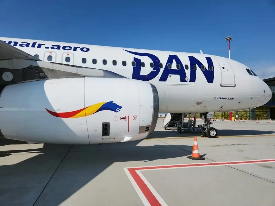 Un avion alb cu inscripția DAN și un motor mare cu un logo colorat sunt parcate pe o pista de beton sub cerul senin.