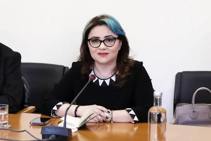 O femeie cu ochelari și păr negru cu reflexii albastre stă la o masă de lemn lângă un microfon și un caiet deschis.