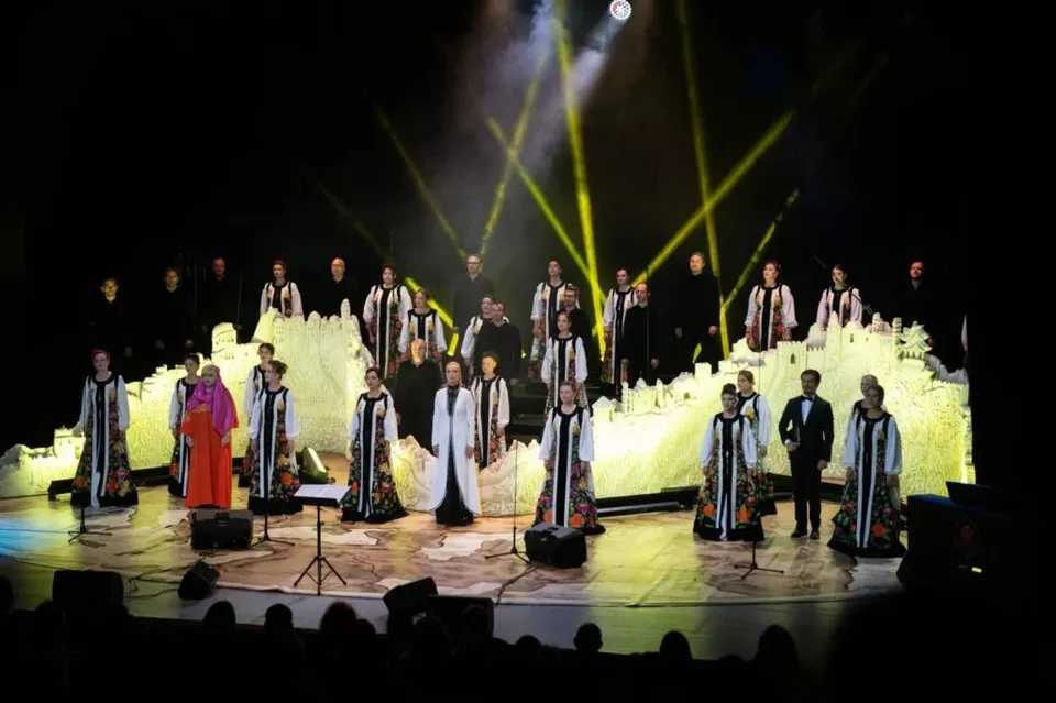 Un grup de persoane în haine tradiționale albe și negre cântă pe o scenă iluminată de lumini galbene sub un fundal de structuri albe luminoase.