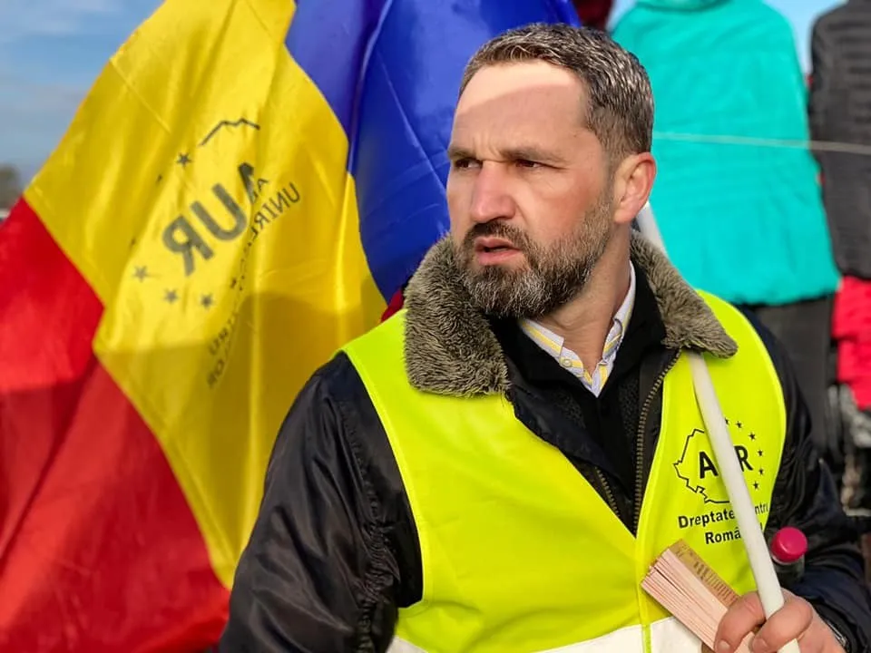 Un bărbat cu barbă poartă o vestă reflectorizantă galbenă și ține în mână un obiect subțire și un dosar în fața unor steaguri colorate.