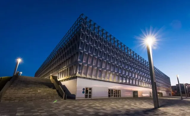 O clădire modernă cu fațadă geometrică iluminată este vizibilă sub un cer albastru, lângă o scară exterioară și un stâlp de iluminat.