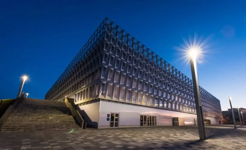 O clădire modernă cu fațadă geometrică iluminată este vizibilă sub un cer albastru, lângă o scară exterioară și un stâlp de iluminat.