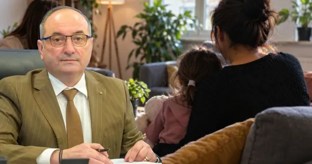 Un bărbat cu ochelari și sacou maro stă la o masă, în timp ce în fundal două femei stau pe o canapea lângă plante de interior.