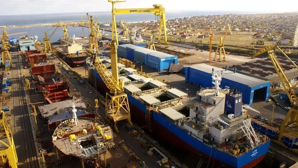 O vedere aeriană a unui șantier naval cu nave mari, macarale galbene și diverse hale industriale de culoare albastră și gri.