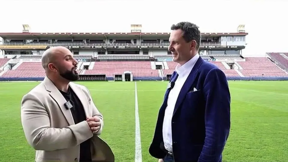 Un bărbat cu barbă poartă un sacou deschis și o cămașă neagră, stând pe un teren de fotbal verde lângă un alt bărbat în sacou albastru.