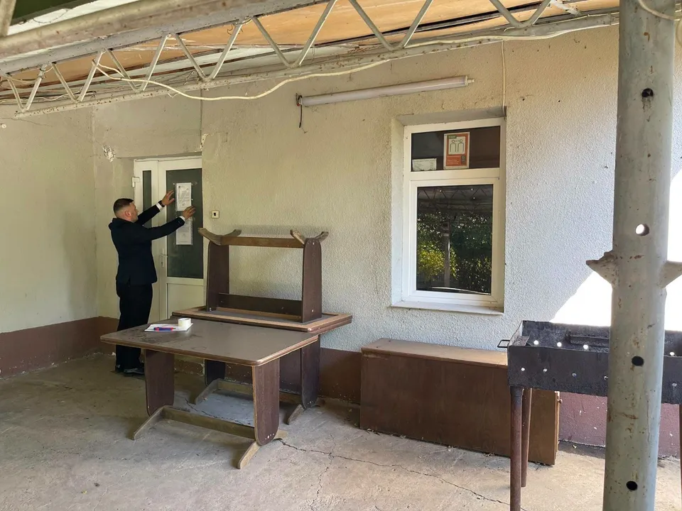 Un bărbat într-un costum închis afișează o foaie albă pe o ușă de sticlă într-o încăpere cu mobilier și o fereastră.