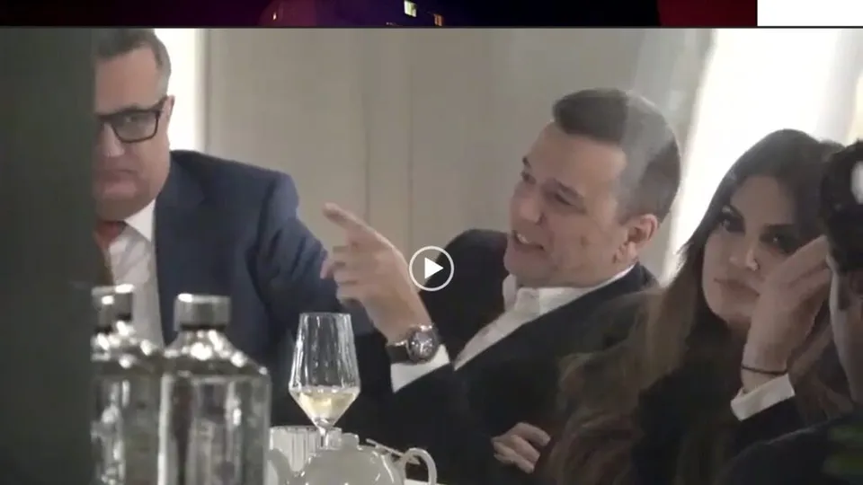 Patru persoane stau la o masă cu pahare și sticle de apă, un bărbat arătând spre stânga în timp ce vorbește.