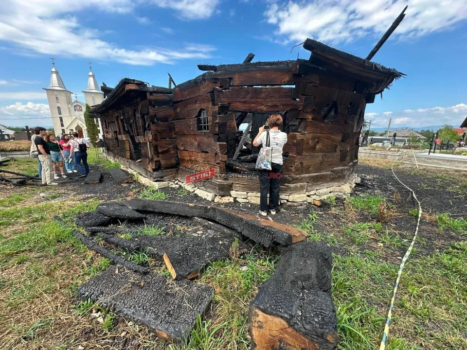 O persoană cu o geantă albă fotografiază structura unei clădiri din lemn carbonizate, înconjurate de resturi de lemn ars pe un teren cu iarbă.