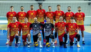 O echipă de volei în echipamente roșii și galbene pozează pe un teren albastru, cu unii jucători în picioare și alții în genunchi.