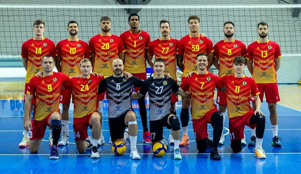 O echipă de volei în echipamente roșii și galbene pozează pe un teren albastru, cu unii jucători în picioare și alții în genunchi.