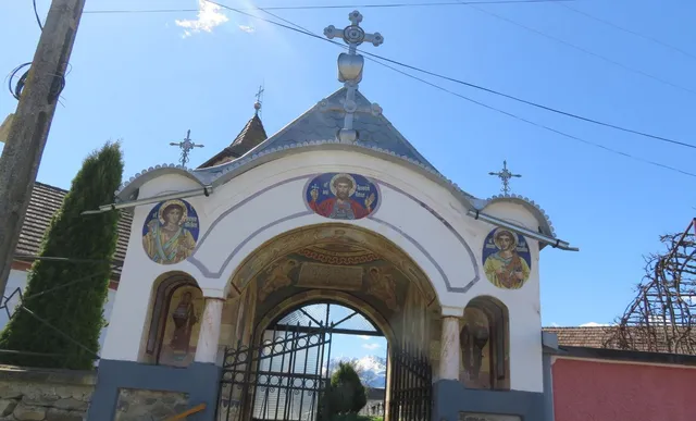 O poartă albă cu arcuri este decorată cu trei picturi religioase circulare și este încoronată de cruci metalice sub un cer senin.