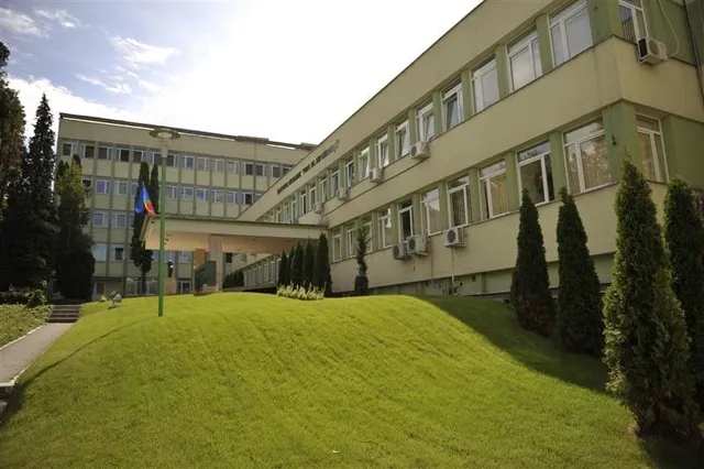 O clădire lungă de culoare deschisă cu ferestre multiple este situată în spatele unui teren de iarbă verde și a unor copaci înalți.