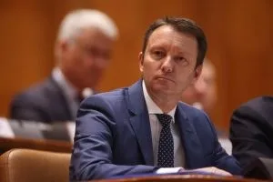 Un bărbat cu părul scurt și costum albastru stă așezat la o masă în fața unui fundal de lemn maro.