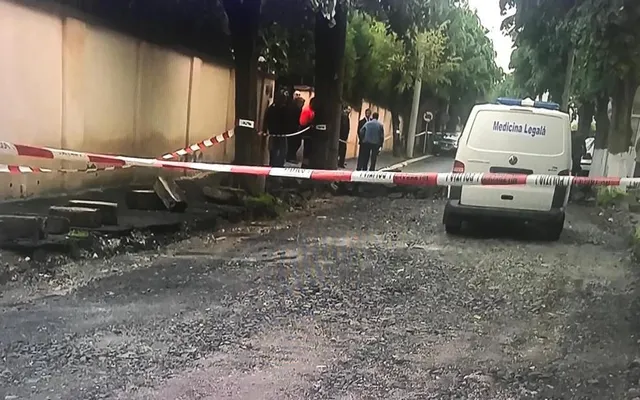 O mașină albă cu inscripția Medicina Legală este parcată pe un drum de pietriș lângă un cordon de poliție din benzi roșii și albe.