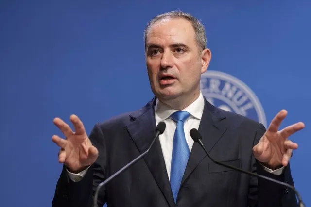 Un bărbat cu părul scurt, purtând sacou închis la culoare, cămașă albă și cravată albastră, stă în fața a două microfoane cu mâinile ridicate.