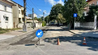 O stradă cu asfalt deteriorat prezintă conuri de circulație portocalii și un semn albastru cu o săgeată albă orientată spre stânga jos.