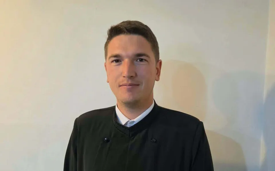 Un bărbat cu părul scurt și haine negre stă în fața unui perete alb.