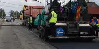 Un utilaj de asfaltare și un camion galben stau pe un drum, în timp ce lucrători în veste reflectorizante execută lucrări.