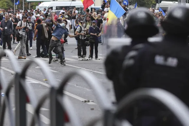 O mulțime de oameni stă pe o stradă lângă polițiști în uniformă, în timp ce un bărbat în cămașă albastră ridică brațele în sus.