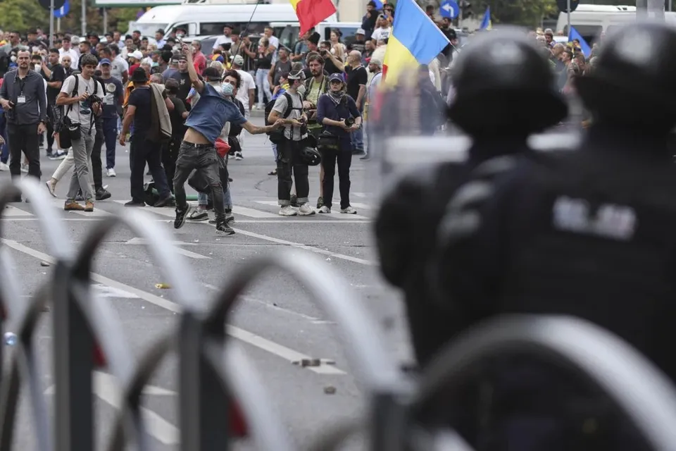 O mulțime de oameni stă pe o stradă lângă polițiști în uniformă, în timp ce un bărbat în cămașă albastră ridică brațele în sus.