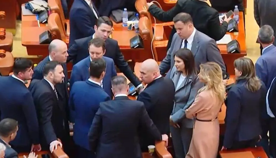 Un grup de oameni îmbrăcați în costume și haine office stau adunați într-o sală cu scaune de lemn și mese maronii.