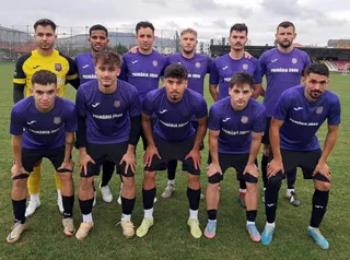 O echipă de fotbaliști în echipamente albastre și galben purtând șosete negre stau așezați și în picioare pe un teren de iarbă verde.