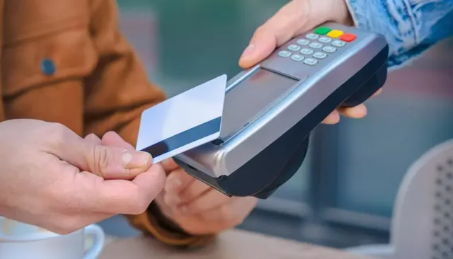 O persoană ține un card bancar alb lângă un terminal de plată de culoare gri și neagră ținut de o altă persoană.