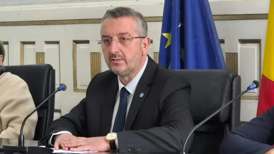 Un bărbat cu ochelari și costum închis la culoare stă la o masă lângă microfoane și steaguri.