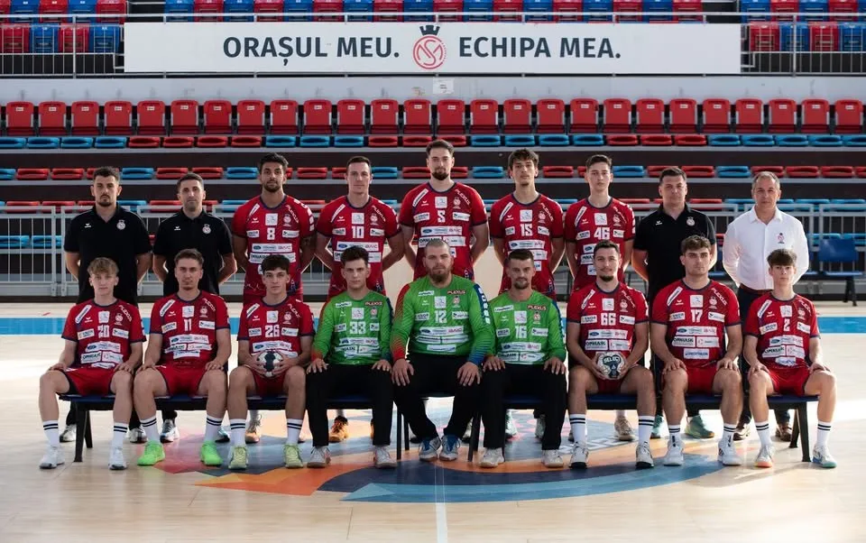 O echipă de handbal masculini poartă echipamente de sport roșii și verzi, așezată în două rânduri în fața unor scaune pe un teren de sport.
