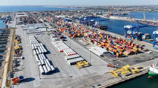 O vedere aeriană a unui port cu numeroase containere colorate, macarale portuare albastre și rulote albe dispuse pe un teren asfaltat lângă apă.