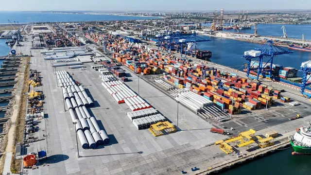 O vedere aeriană a unui port cu numeroase containere colorate, macarale portuare albastre și rulote albe dispuse pe un teren asfaltat lângă apă.