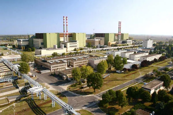 O vedere aeriană prezintă un complex industrial cu clădiri mari, conducte albe și două turnuri înalte cu dungi roșii și albe sub un cer senin.