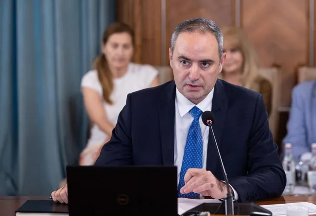 Un bărbat într-un costum închis la culoare și cravată albastră vorbește la un microfon în fața unui laptop.