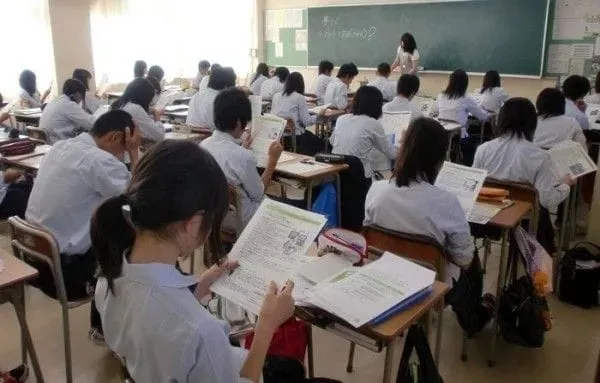 Elevi în uniforme albe stau la bănci într-o sală de clasă și lucrează cu manuale și caiete deschise.