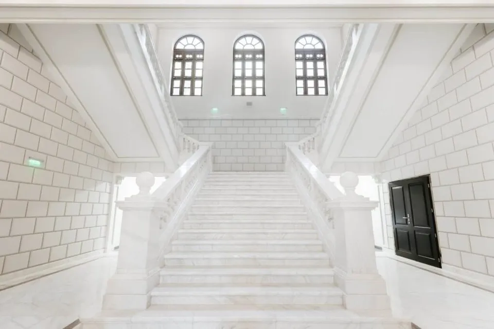 O scară interioară de marmură albă cu balustrade masive se ridică spre ferestre arcuite într-o încăpere cu pereți albi texturați.