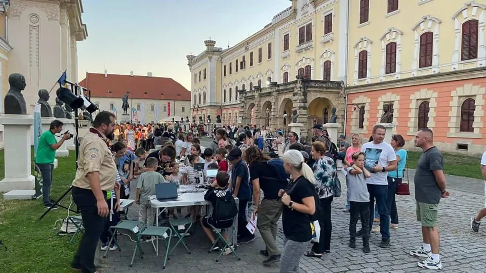 Un grup de oameni stă la mese albe în fața unor clădiri mari, participând la activități în aer liber pe un teren cu iarbă și pietriș.