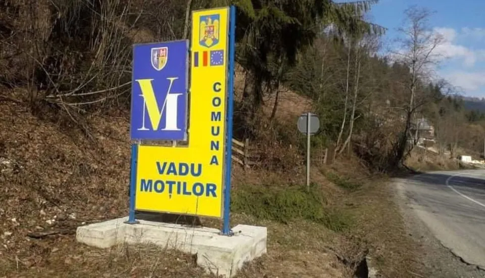 Un panou informativ galben și albastru cu textul Vadu Moților este amplasat pe marginea unui drum lângă o pădure.