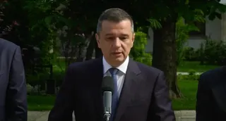 Un bărbat cu părul scurt și costum închis, purtând cămașă albă și cravată albastră, vorbește în fața unui microfon aflat în prim-plan.