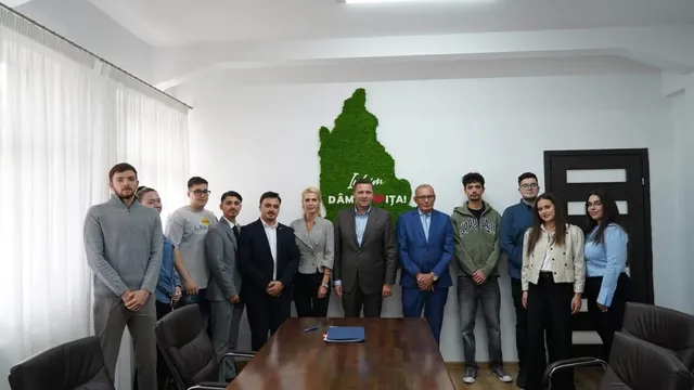 Un grup de oameni stă în picioare într-o încăpere lângă o masă de lemn, în fața unui perete alb cu o formă verde.