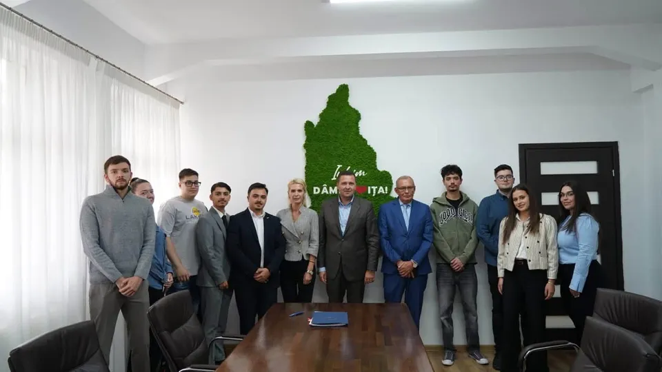 Un grup de oameni stă în picioare într-o încăpere lângă o masă de lemn, în fața unui perete alb cu o formă verde.